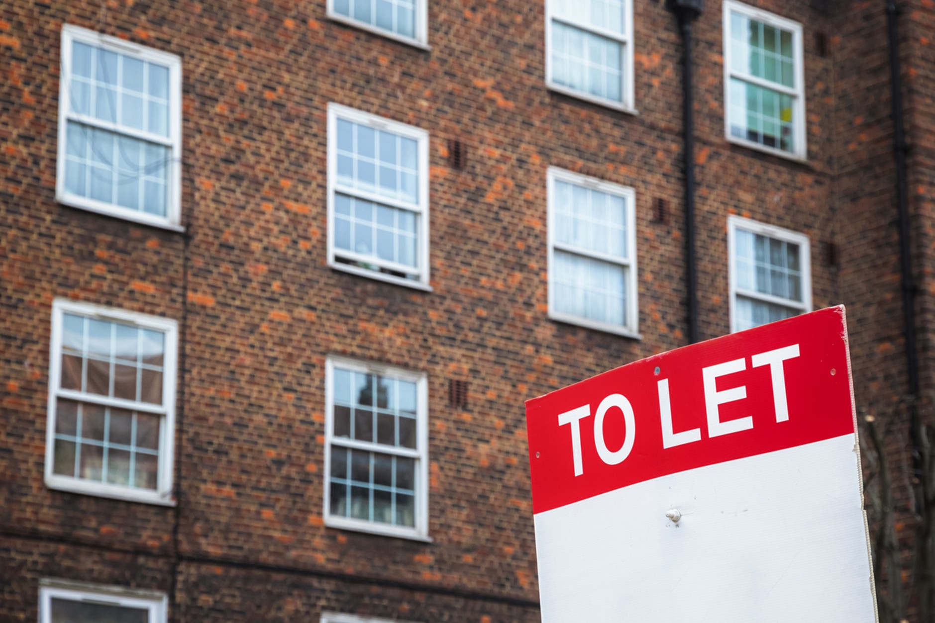 A red and white “To Let” sign in front of a brick apartment building with multiple windows.
