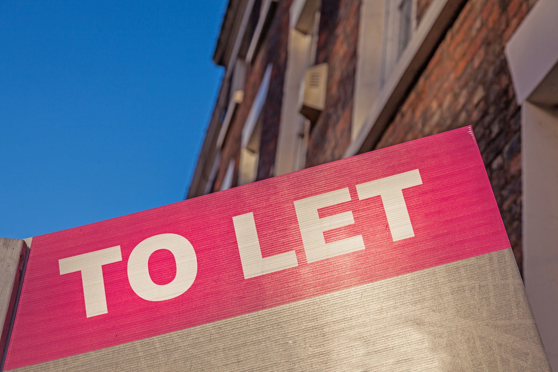 Close-up of a pink sign with large white letters reading “TO LET,” set against a brick building and blue sky in the background.