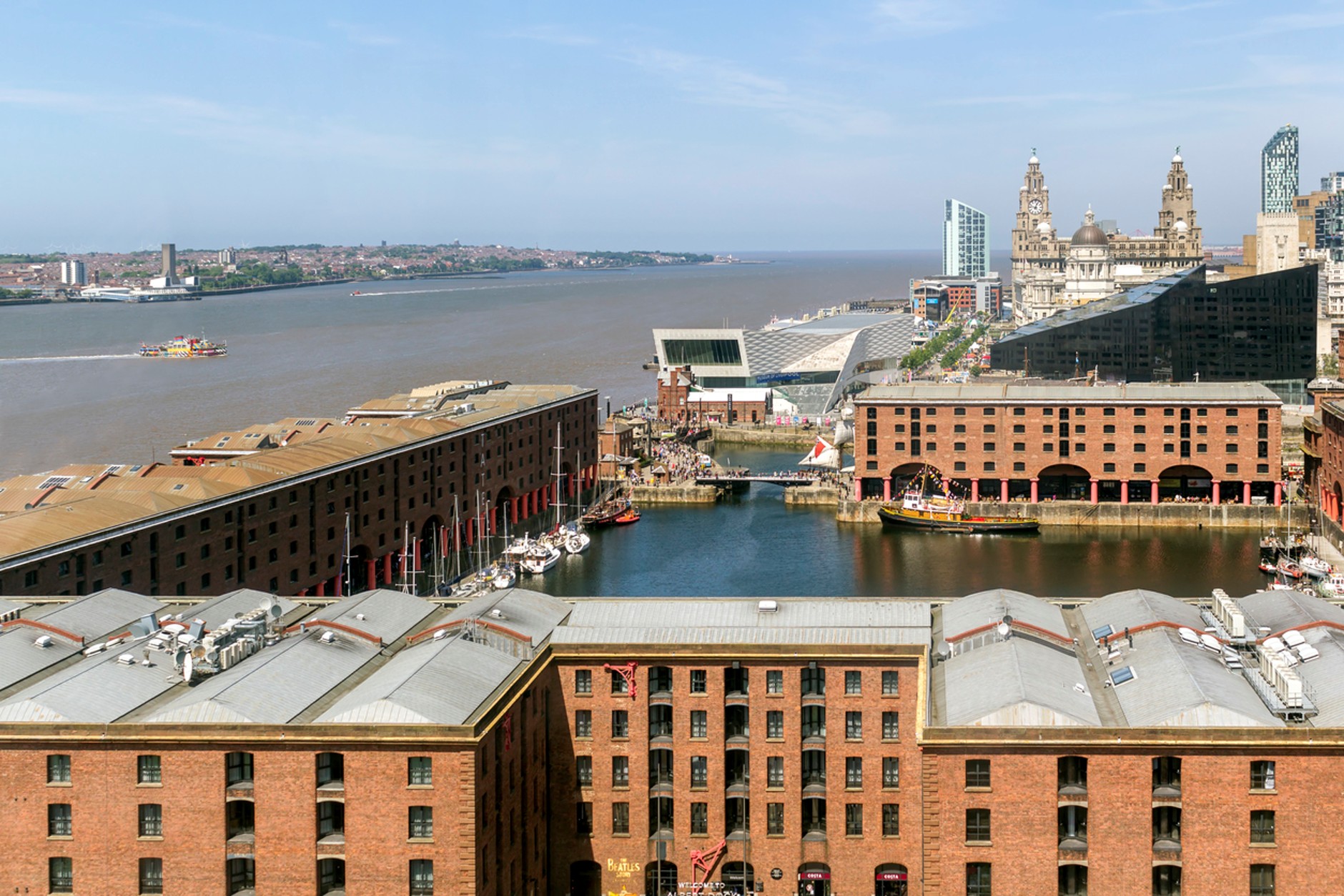 View over Liverpool's historic Albert Dock and waterfront on a clear day, showing red-brick dock buildings, moored boats, the River Mersey, and the city skyline including the Royal Liver Building in the background.