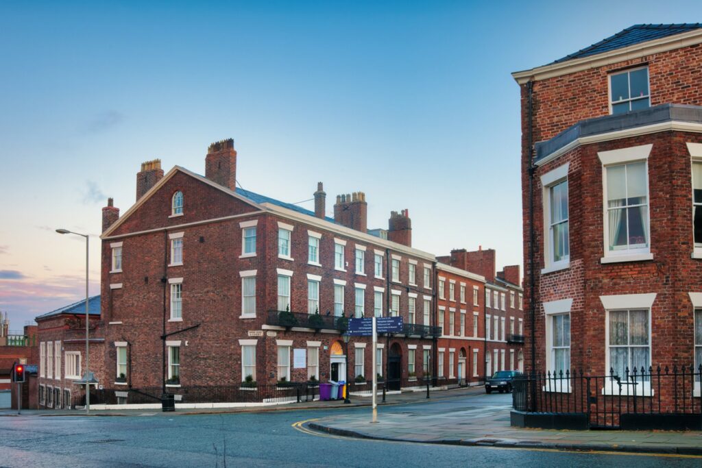 Historic red-brick Georgian buildings lining a quiet street in Liverpool, with traditional sash windows, chimneys, and a clear blue sky overhead.