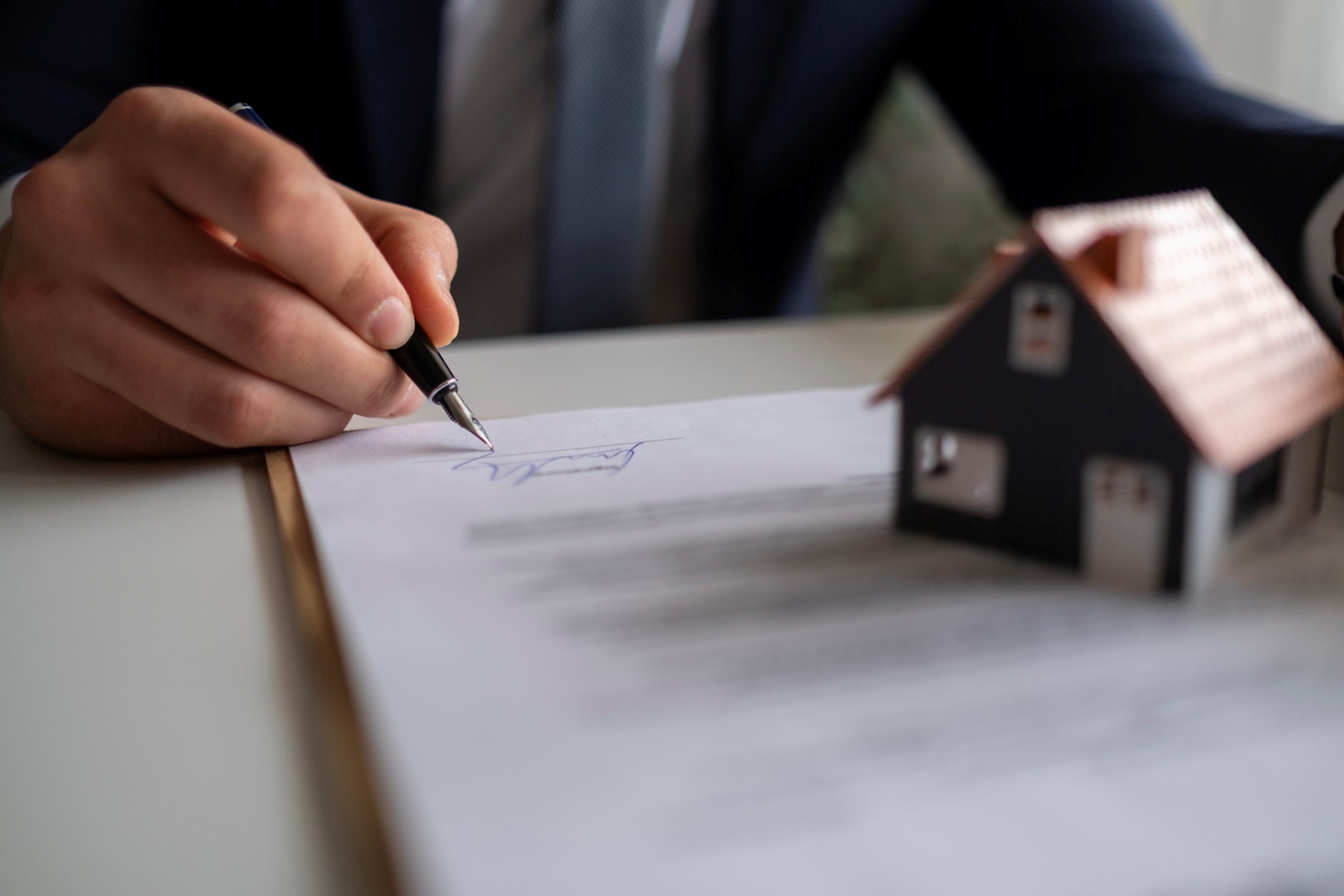 Close-up of a person signing a property-related document with a pen, with a small model house in the foreground, representing a home purchase, sale, or rental agreement.