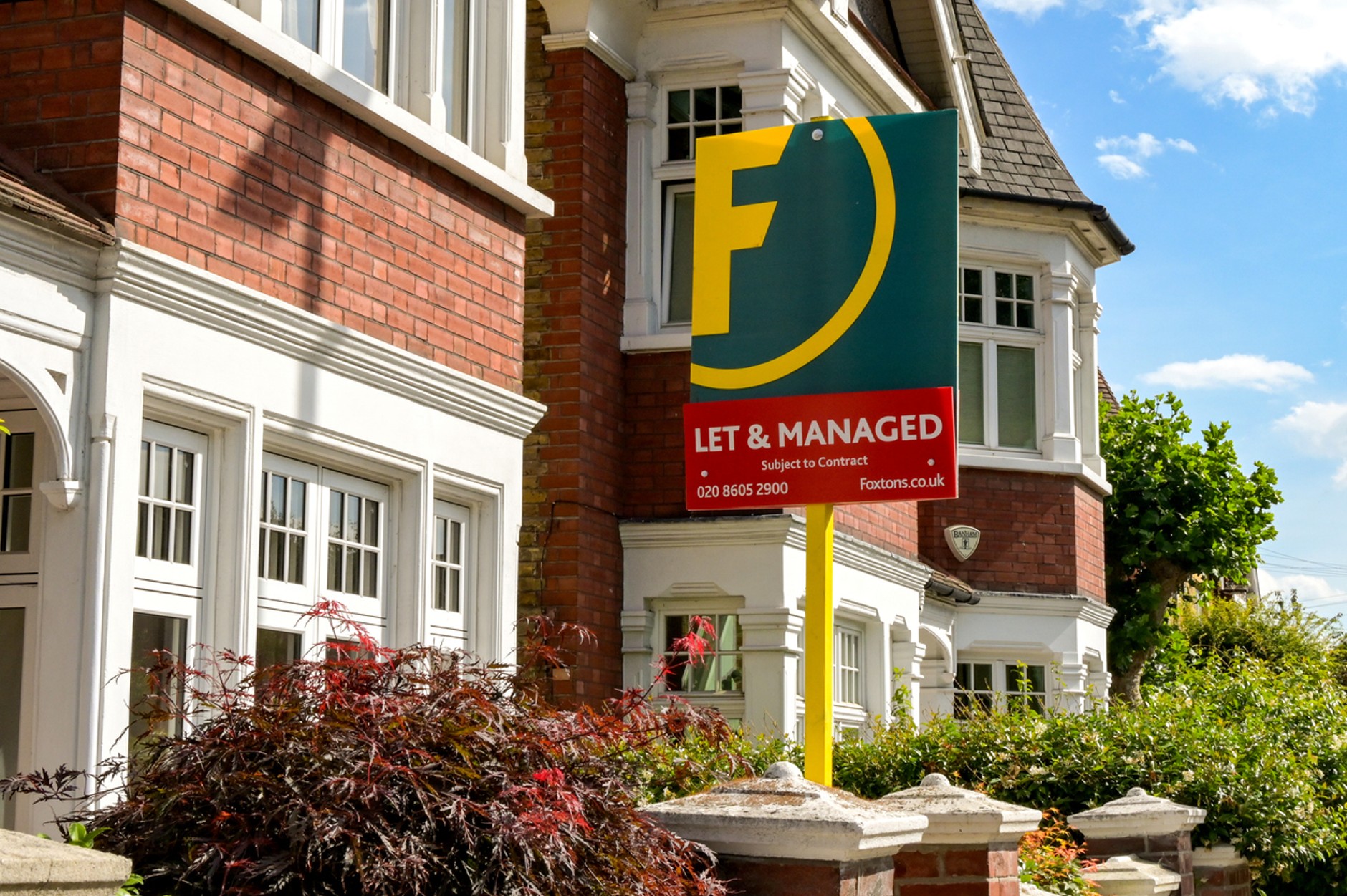 Foxtons “Let & Managed” estate agent sign displayed outside a large red-brick residential property with white-framed windows, surrounded by shrubs and greenery on a sunny day.