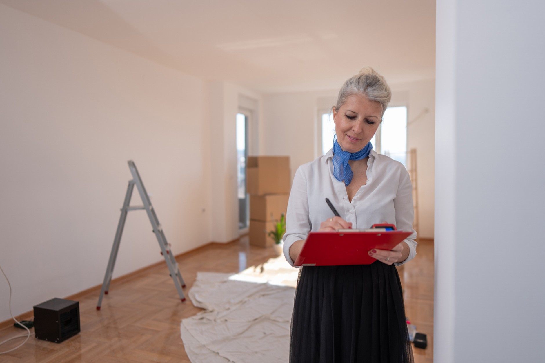 Person carrying out a property inspection in an empty room, writing notes on a clipboard. The room features wooden flooring, a step ladder, moving boxes, and protective floor coverings, suggesting preparation for sale, letting, or renovation.