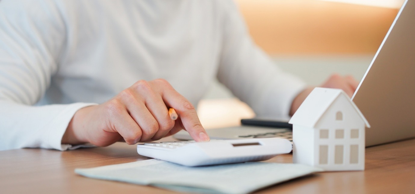 Person using a calculator at a desk beside a laptop and a miniature house model, representing property finance, mortgage calculations, or home-buying costs.