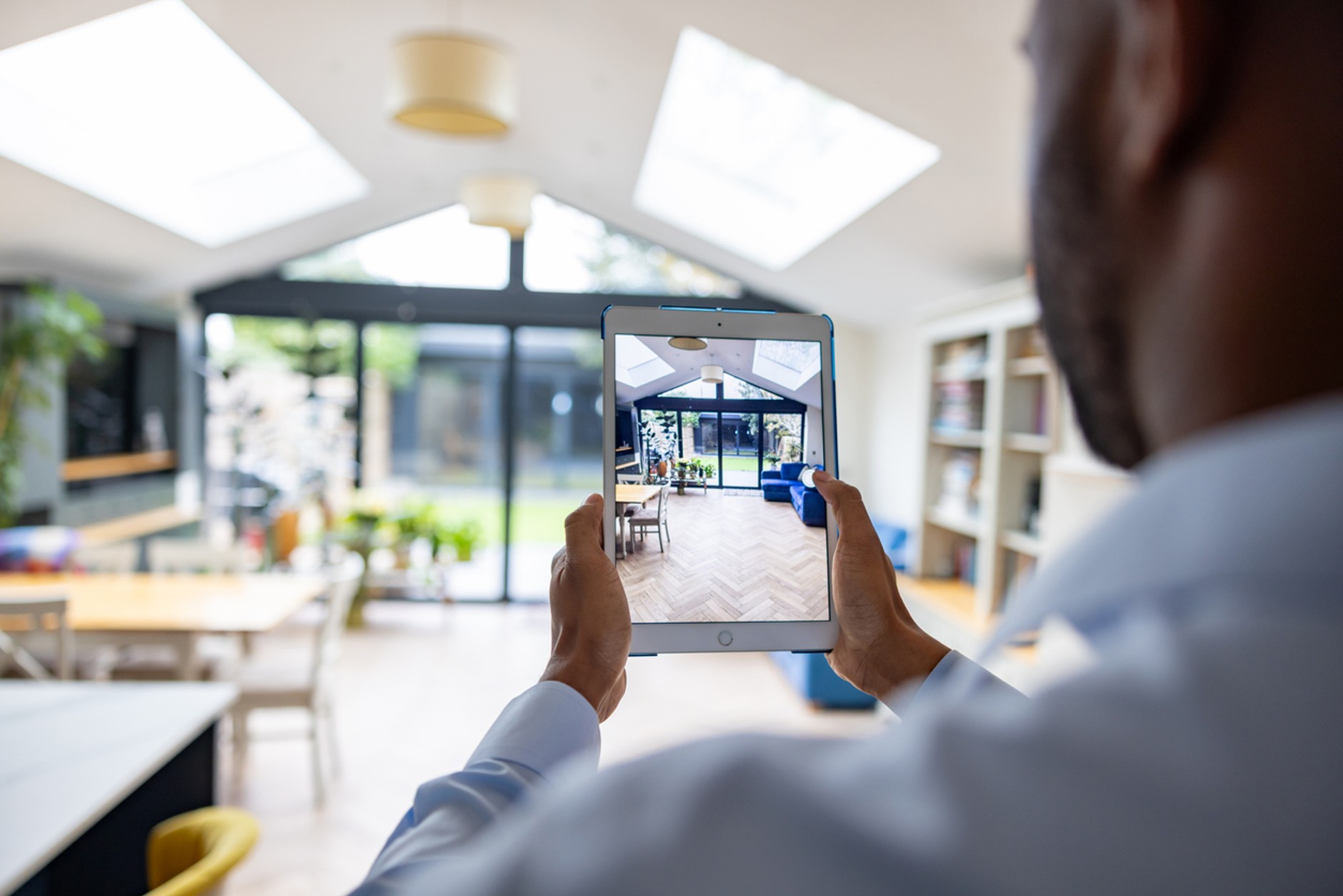 Person using a tablet to photograph or create a virtual tour of a bright, modern open-plan living space with large skylights, floor-to-ceiling glass doors, wooden flooring, and contemporary furnishings.