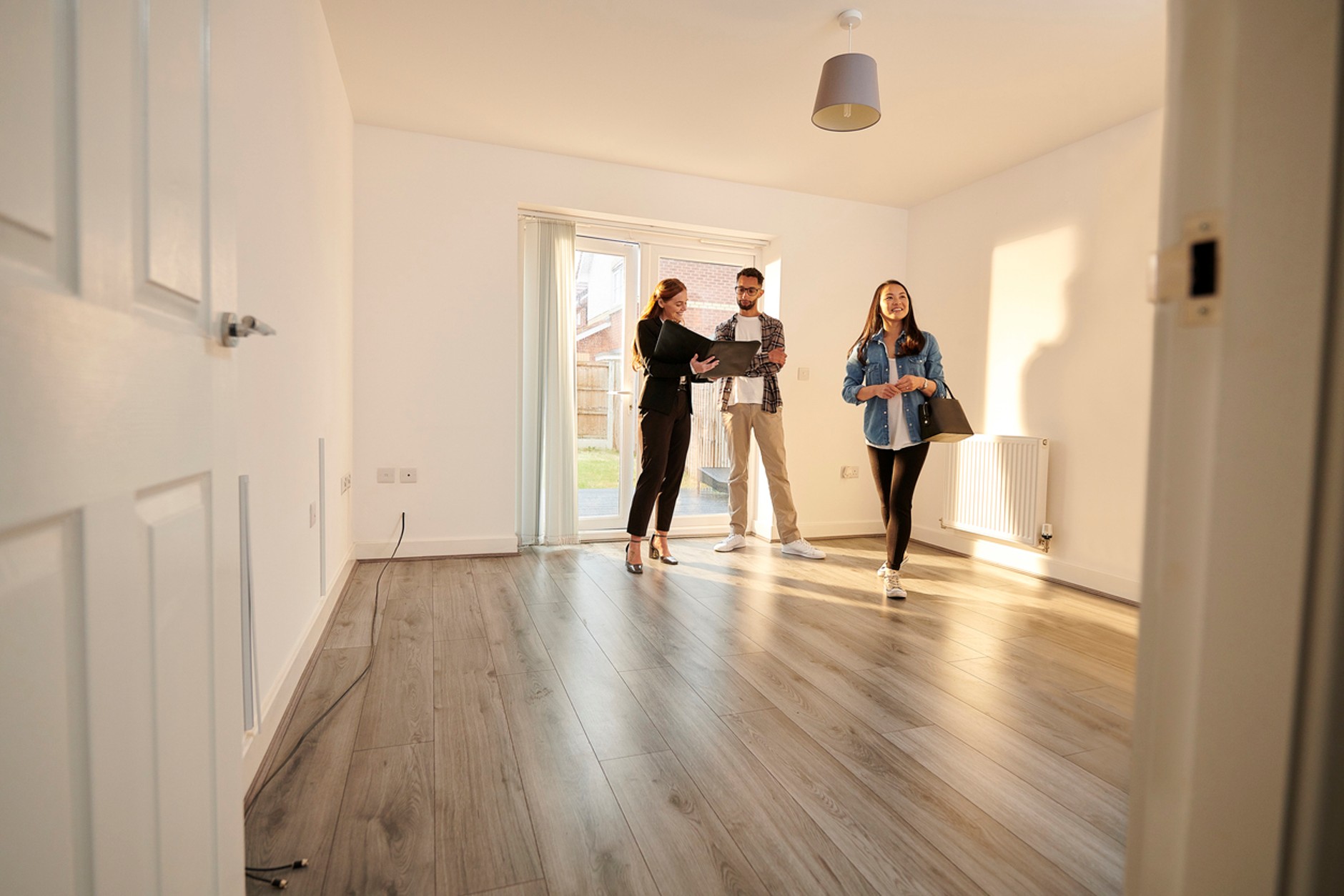 Prospective tenants or buyers viewing an empty property with an estate agent. The bright room features wood-effect flooring, white walls, a radiator, and patio doors leading to a garden, while the agent discusses details from a property folder.