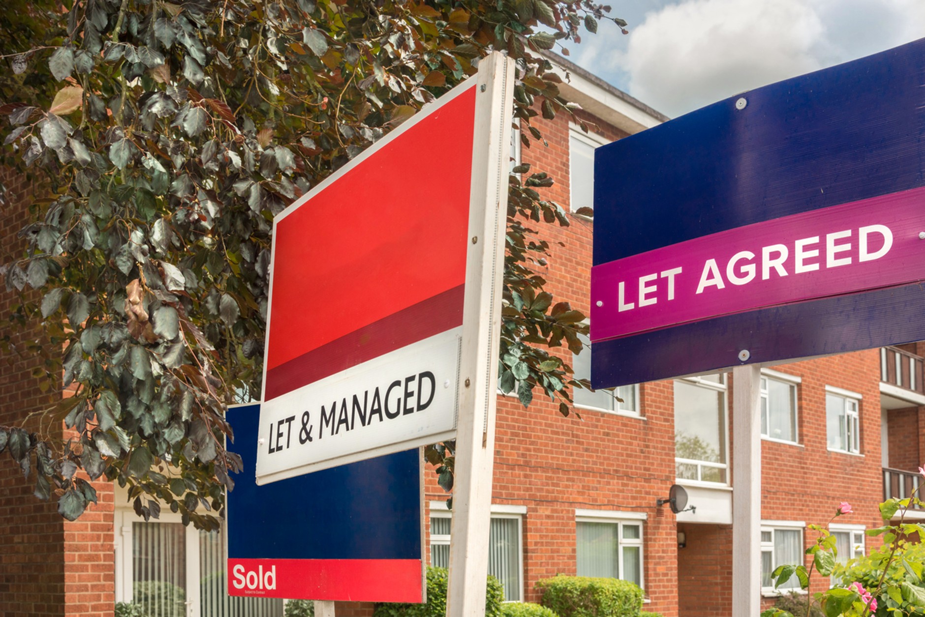 Residential property marketing boards outside a block of flats, including signs reading “Let & Managed,” “Let Agreed,” and “Sold,” indicating successful property sales and lettings. Trees and a red-brick apartment building are visible in the background.